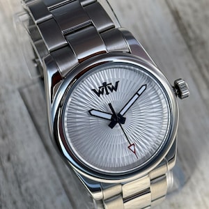 Puede incluir: Un reloj de pulsera de acero inoxidable plateado con una correa de eslabones met&aacute;licos y una esfera redonda con textura de rayos de sol. La esfera muestra el texto negro WTW en la parte superior, con manecillas de borde negro y un segundero con punta roja.