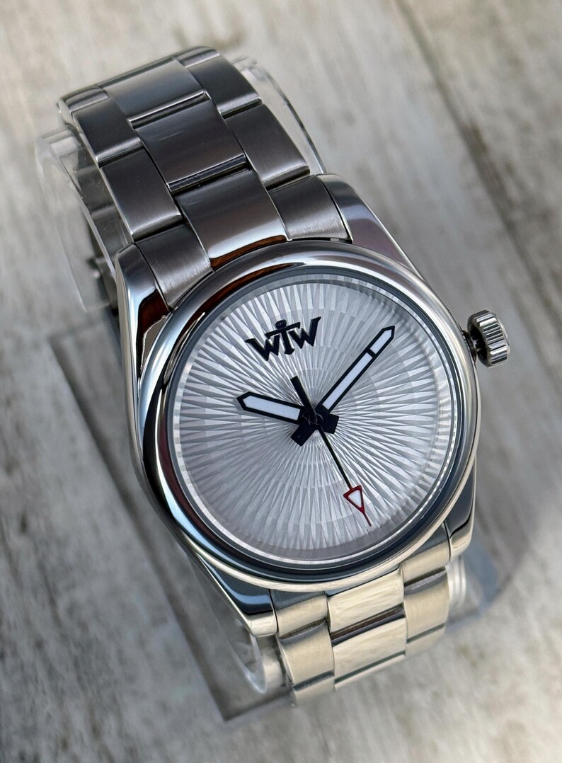 Puede incluir: Un reloj de pulsera de acero inoxidable plateado con una correa de eslabones met&aacute;licos y una esfera redonda con textura de rayos de sol. La esfera muestra el texto negro WTW en la parte superior, con manecillas de borde negro y un segundero con punta roja.