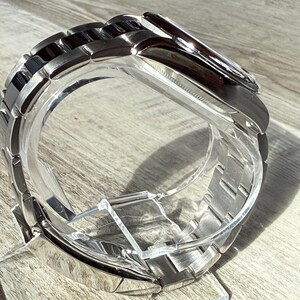 Puede incluir: Un reloj de pulsera con una correa de eslabones de metal plateado pulido con eslabones centrales negros. La caja del reloj es redonda con un bisel plateado y una esfera oscura, presentado sobre un soporte de acr&iacute;lico transparente.