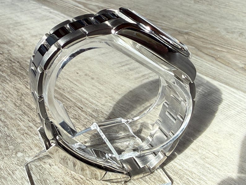 Puede incluir: Un reloj de pulsera con una correa de eslabones de metal plateado pulido con eslabones centrales negros. La caja del reloj es redonda con un bisel plateado y una esfera oscura, presentado sobre un soporte de acr&iacute;lico transparente.