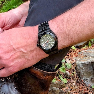 K&ouml;nnte beinhalten: Eine schwarze Armbanduhr mit mattem Finish und einem strukturierten, geometrisch gemusterten Zifferblatt in Grau- und Silbert&ouml;nen. Die Uhr verf&uuml;gt &uuml;ber ein schwarzes Metallgliederarmband, eine runde L&uuml;nette mit sichtbaren Schrauben und einen roten Sekundenzeiger.