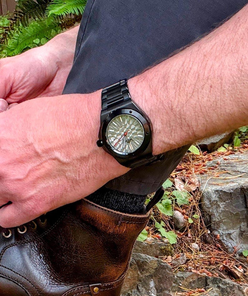 K&ouml;nnte beinhalten: Eine schwarze Armbanduhr mit mattem Finish und einem strukturierten, geometrisch gemusterten Zifferblatt in Grau- und Silbert&ouml;nen. Die Uhr verf&uuml;gt &uuml;ber ein schwarzes Metallgliederarmband, eine runde L&uuml;nette mit sichtbaren Schrauben und einen roten Sekundenzeiger.