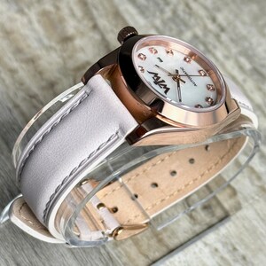May include: An automatic wristwatch with a rose gold-tone metal case and a white leather strap. The watch face features a white mother-of-pearl dial with small crystal hour markers, rose gold hands, and the black text W7W and AUTOMATIC.
