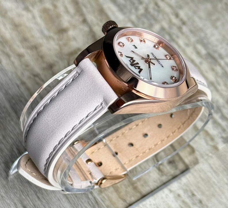 May include: An automatic wristwatch with a rose gold-tone metal case and a white leather strap. The watch face features a white mother-of-pearl dial with small crystal hour markers, rose gold hands, and the black text W7W and AUTOMATIC.