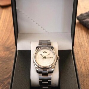 Puede incluir: Un reloj de pulsera plateado con una esfera blanca de patr&oacute;n radial y las letras WtW impresas en negro. El reloj cuenta con una correa de eslabones met&aacute;licos y se presenta dentro de una caja de presentaci&oacute;n rectangular negra abierta.