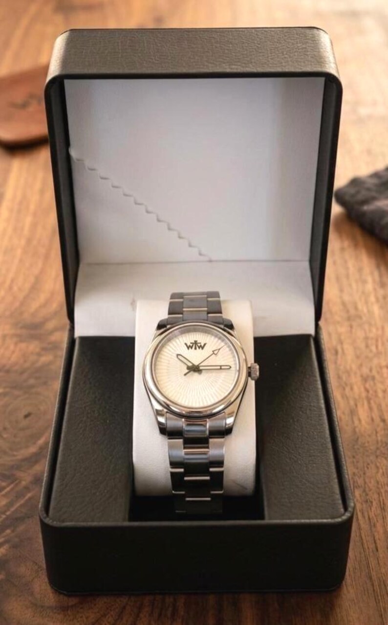 Puede incluir: Un reloj de pulsera plateado con una esfera blanca de patr&oacute;n radial y las letras WtW impresas en negro. El reloj cuenta con una correa de eslabones met&aacute;licos y se presenta dentro de una caja de presentaci&oacute;n rectangular negra abierta.