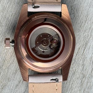 May include: A rose gold-toned watch case with a clear exhibition case back revealing the internal mechanical movement. The watch features a metallic crown on the left side and is attached to a light beige leather strap with visible stitching.