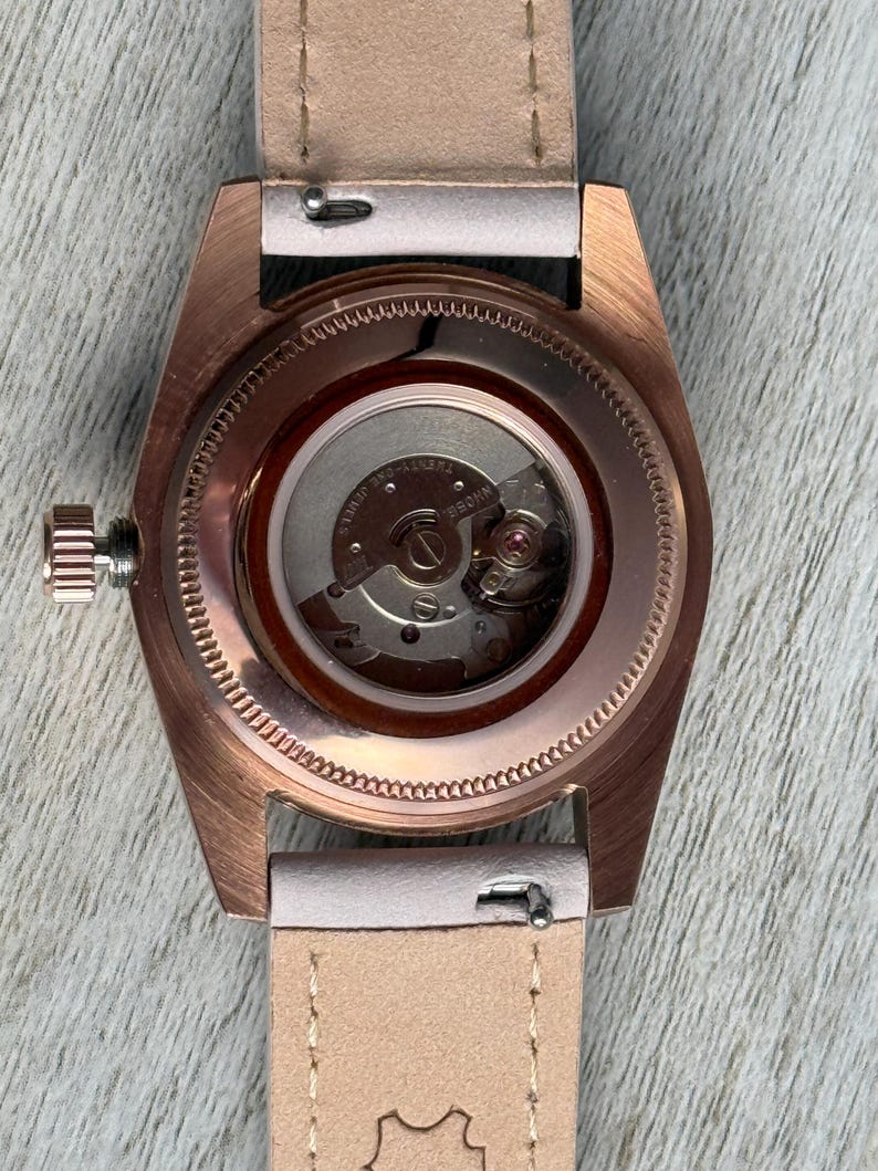 May include: A rose gold-toned watch case with a clear exhibition case back revealing the internal mechanical movement. The watch features a metallic crown on the left side and is attached to a light beige leather strap with visible stitching.