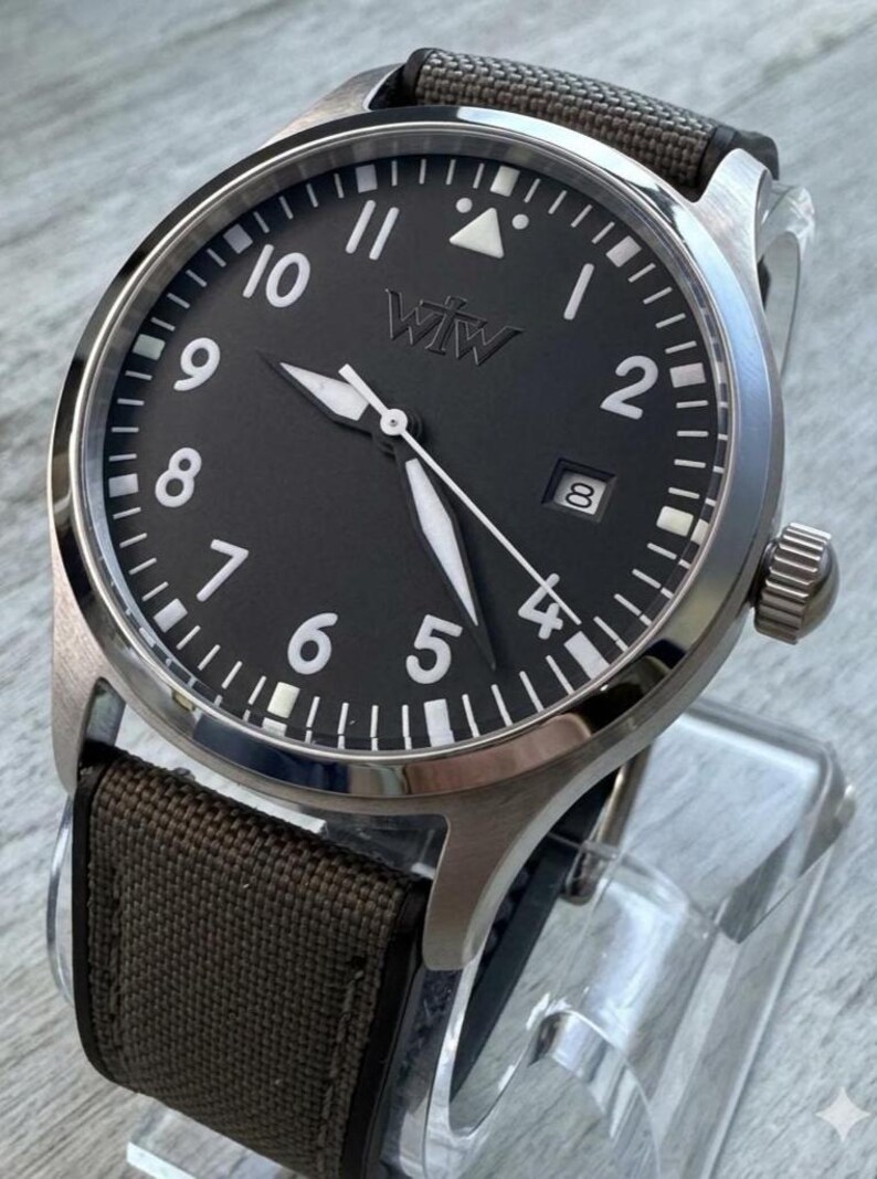 K&ouml;nnte beinhalten: Eine Armbanduhr mit rundem, silberfarbenem Metallgeh&auml;use und mattschwarzem Zifferblatt. Das Zifferblatt zeigt wei&szlig;e arabische Ziffern, ein Datumsfenster auf der Drei-Uhr-Position und den Schriftzug "WTW" unter der Zw&ouml;lf-Uhr-Markierung. Braunes Textilarmband.