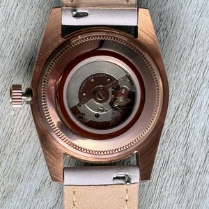 May include: A rose gold-toned wristwatch with an exhibition case back revealing the internal mechanical movement. The watch features a light beige leather strap with visible stitching and a side-mounted winding crown on the left side of the case.