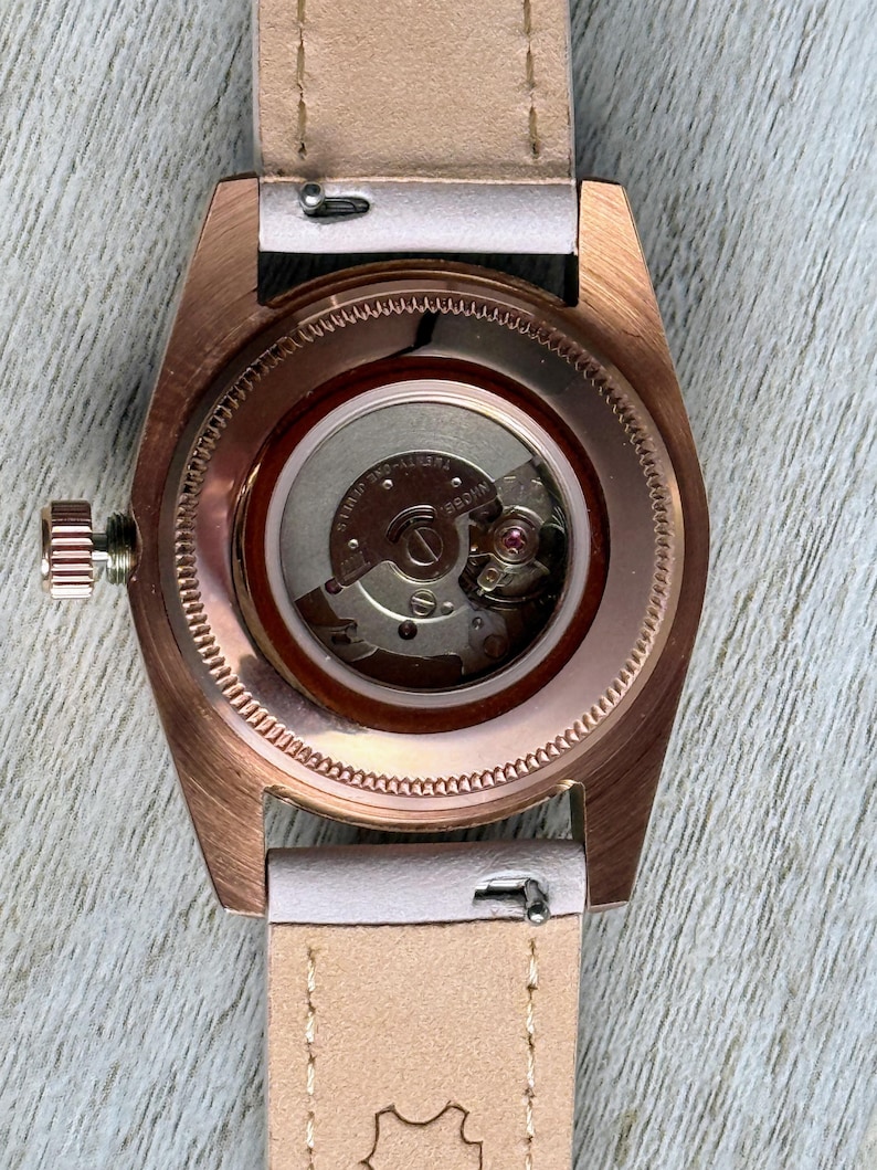 May include: A rose gold-toned wristwatch with an exhibition case back revealing the internal mechanical movement. The watch features a light beige leather strap with visible stitching and a side-mounted winding crown on the left side of the case.