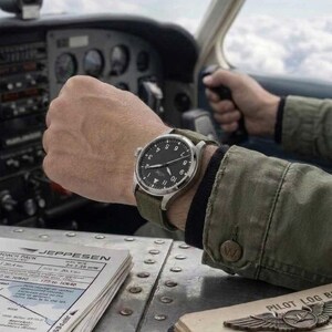 K&ouml;nnte beinhalten: Eine Armbanduhr aus Edelstahl mit schwarzem Zifferblatt, wei&szlig;en arabischen Ziffern und einem gr&uuml;nen Canvas-Armband. Die Uhr hat ein rundes Geh&auml;use und wird am Handgelenk in einem Flugzeugcockpit getragen, neben einer Jeppesen-Karte und einem Flugbuch.