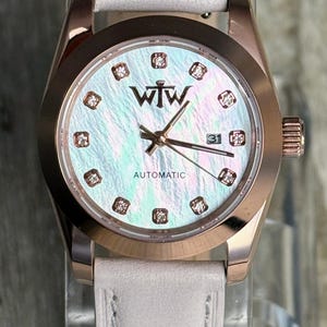 May include: An automatic wristwatch with a rose gold-tone case and a light grey leather strap. The iridescent mother-of-pearl dial features the WTW logo, diamond-accented hour markers, rose gold hands, and a date window displaying the number 31.
