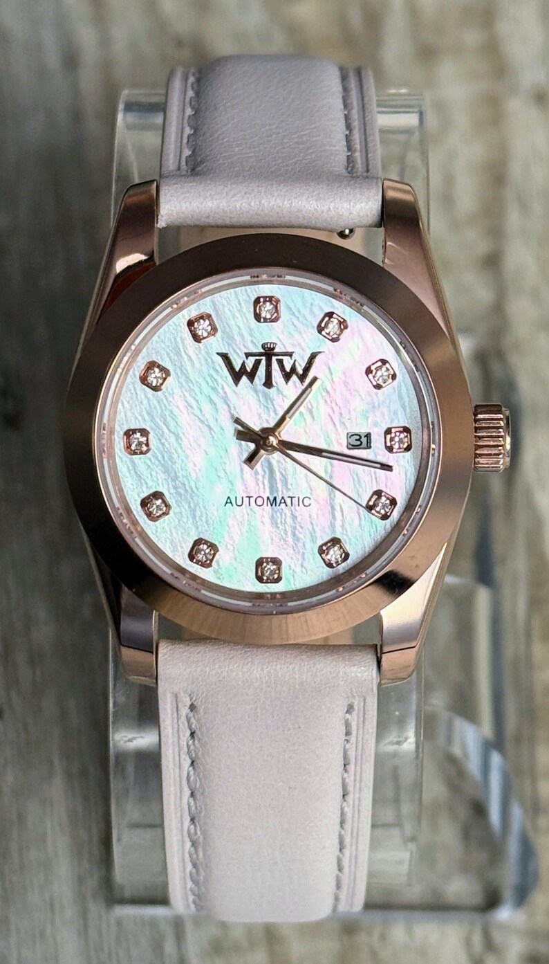 May include: An automatic wristwatch with a rose gold-tone case and a light grey leather strap. The iridescent mother-of-pearl dial features the WTW logo, diamond-accented hour markers, rose gold hands, and a date window displaying the number 31.