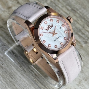 May include: An automatic wristwatch with a rose gold-tone case and a light pink leather strap. The white mother-of-pearl dial features rose gold hands, a date window at the three o'clock position, and crystal hour markers with the WTW logo.