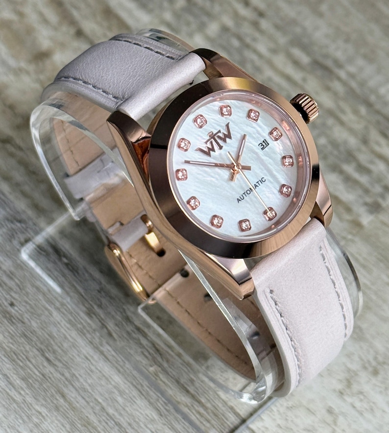 May include: An automatic wristwatch with a rose gold-tone case and a light pink leather strap. The white mother-of-pearl dial features rose gold hands, a date window at the three o'clock position, and crystal hour markers with the WTW logo.