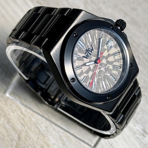 K&ouml;nnte beinhalten: Eine automatische Armbanduhr aus schwarzem Edelstahl mit einem strukturierten, geometrischen grauen Zifferblatt. Die Uhr hat eine schwarze L&uuml;nette mit vier sichtbaren Schrauben, silberne Stundenmarkierungen, einen roten Sekundenzeiger und ein Datumsfenster mit der Zahl 21.