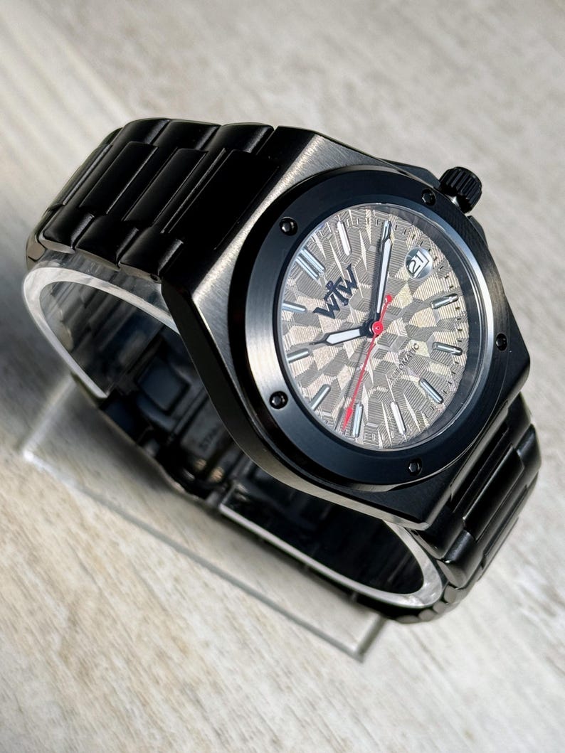 K&ouml;nnte beinhalten: Eine automatische Armbanduhr aus schwarzem Edelstahl mit einem strukturierten, geometrischen grauen Zifferblatt. Die Uhr hat eine schwarze L&uuml;nette mit vier sichtbaren Schrauben, silberne Stundenmarkierungen, einen roten Sekundenzeiger und ein Datumsfenster mit der Zahl 21.