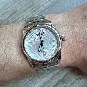 Puede incluir: Un reloj de pulsera plateado con una correa de eslabones met&aacute;licos y una esfera redonda con un patr&oacute;n de rayos de sol. La esfera muestra un logotipo negro en la parte superior, manecillas plateadas con centros blancos y un segundero con punta roja.