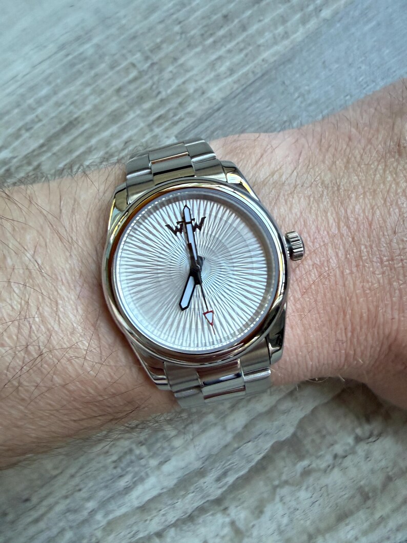 Puede incluir: Un reloj de pulsera plateado con una correa de eslabones met&aacute;licos y una esfera redonda con un patr&oacute;n de rayos de sol. La esfera muestra un logotipo negro en la parte superior, manecillas plateadas con centros blancos y un segundero con punta roja.