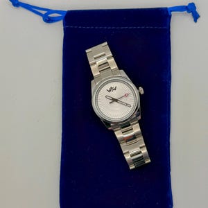 Puede incluir: Un reloj de pulsera de acero inoxidable en tono plateado con una correa de eslabones met&aacute;licos y una esfera redonda con un patr&oacute;n de rayos texturizado. La esfera muestra el logotipo "WfW" en la parte superior, manecillas plateadas y un segundero rojo.