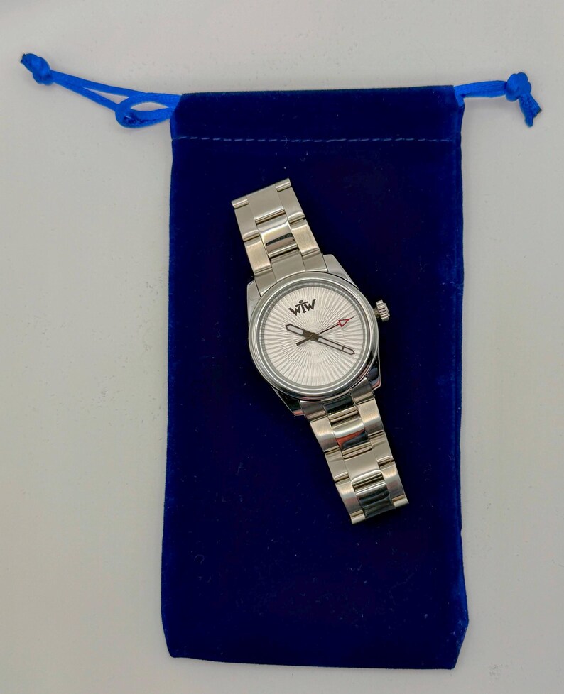 Puede incluir: Un reloj de pulsera de acero inoxidable en tono plateado con una correa de eslabones met&aacute;licos y una esfera redonda con un patr&oacute;n de rayos texturizado. La esfera muestra el logotipo "WfW" en la parte superior, manecillas plateadas y un segundero rojo.
