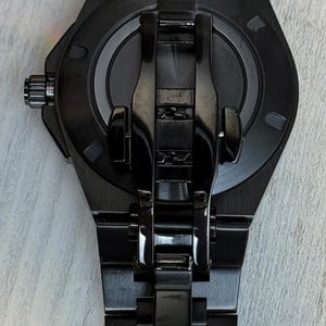 K&ouml;nnte beinhalten: Eine mattschwarze Armbanduhr mit einem runden Geh&auml;use und einem passenden Gliederarmband. Die R&uuml;ckseite der Uhr zeigt eine zentrale kreisf&ouml;rmige Platte mit metallischem Finish, umgeben von einem glatten schwarzen Rahmen und einer Faltschlie&szlig;e.