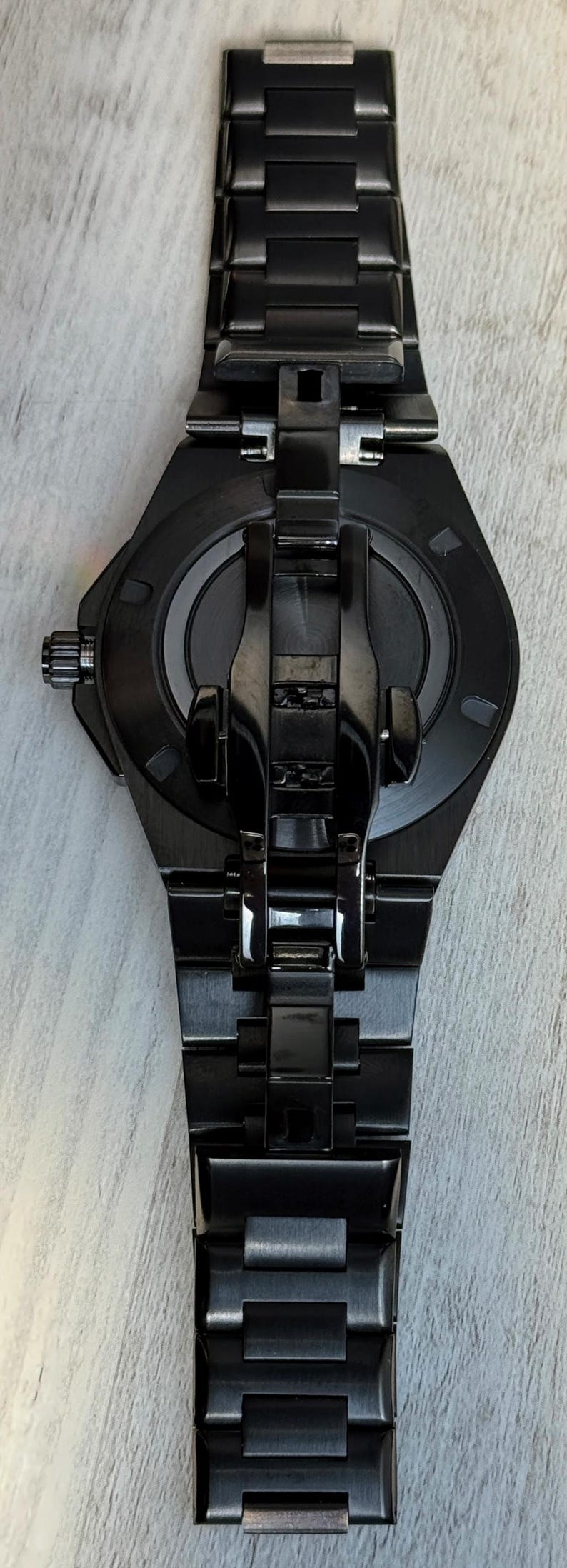 K&ouml;nnte beinhalten: Eine mattschwarze Armbanduhr mit einem runden Geh&auml;use und einem passenden Gliederarmband. Die R&uuml;ckseite der Uhr zeigt eine zentrale kreisf&ouml;rmige Platte mit metallischem Finish, umgeben von einem glatten schwarzen Rahmen und einer Faltschlie&szlig;e.
