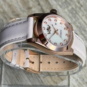 May include: An automatic wristwatch with a rose gold-tone metal case and a white leather strap. The watch face features a white mother-of-pearl dial with rose gold-tone hands, crystal hour markers, and the text "WtW" and "AUTOMATIC" printed on it.