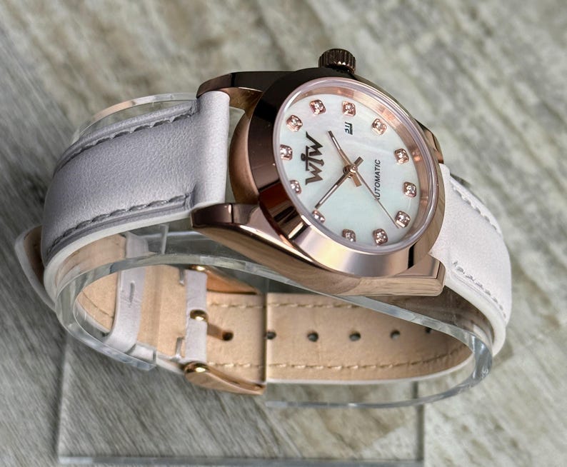 May include: An automatic wristwatch with a rose gold-tone metal case and a white leather strap. The watch face features a white mother-of-pearl dial with rose gold-tone hands, crystal hour markers, and the text "WtW" and "AUTOMATIC" printed on it.