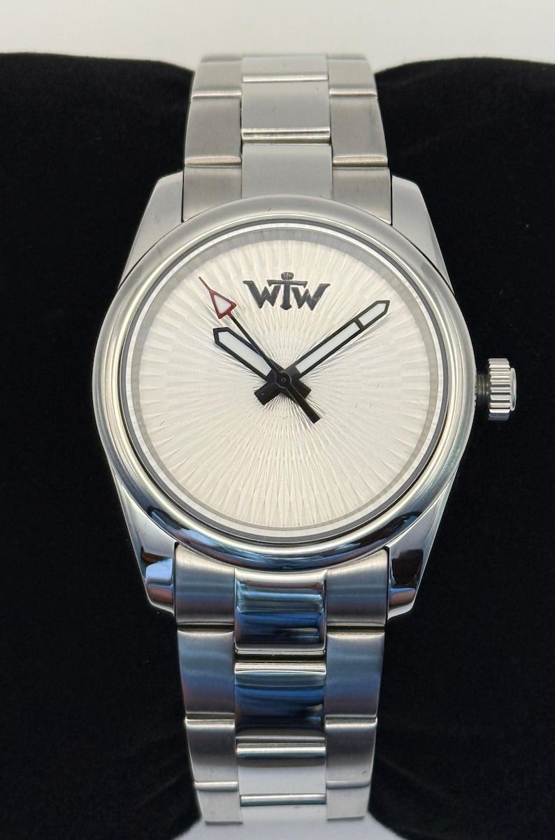 Puede incluir: Un reloj de pulsera de acero inoxidable en tono plateado con una esfera blanca de patr&oacute;n radial y el texto WTW impreso en negro. El reloj incluye una correa de metal de tres eslabones, una corona en el lado derecho y manecillas con bordes negros.