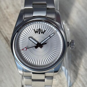 Puede incluir: Un reloj de pulsera de acero inoxidable con esfera plateada de rayos de sol y el logotipo negro WTW. El reloj cuenta con manecillas luminosas, un segundero negro fino con punta triangular roja y una correa de eslabones de metal cepillado.