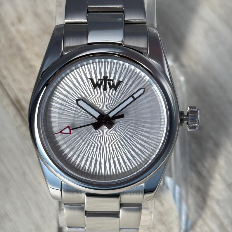 Puede incluir: Un reloj de pulsera de acero inoxidable con esfera plateada de rayos de sol y el logotipo negro WTW. El reloj cuenta con manecillas luminosas, un segundero negro fino con punta triangular roja y una correa de eslabones de metal cepillado.