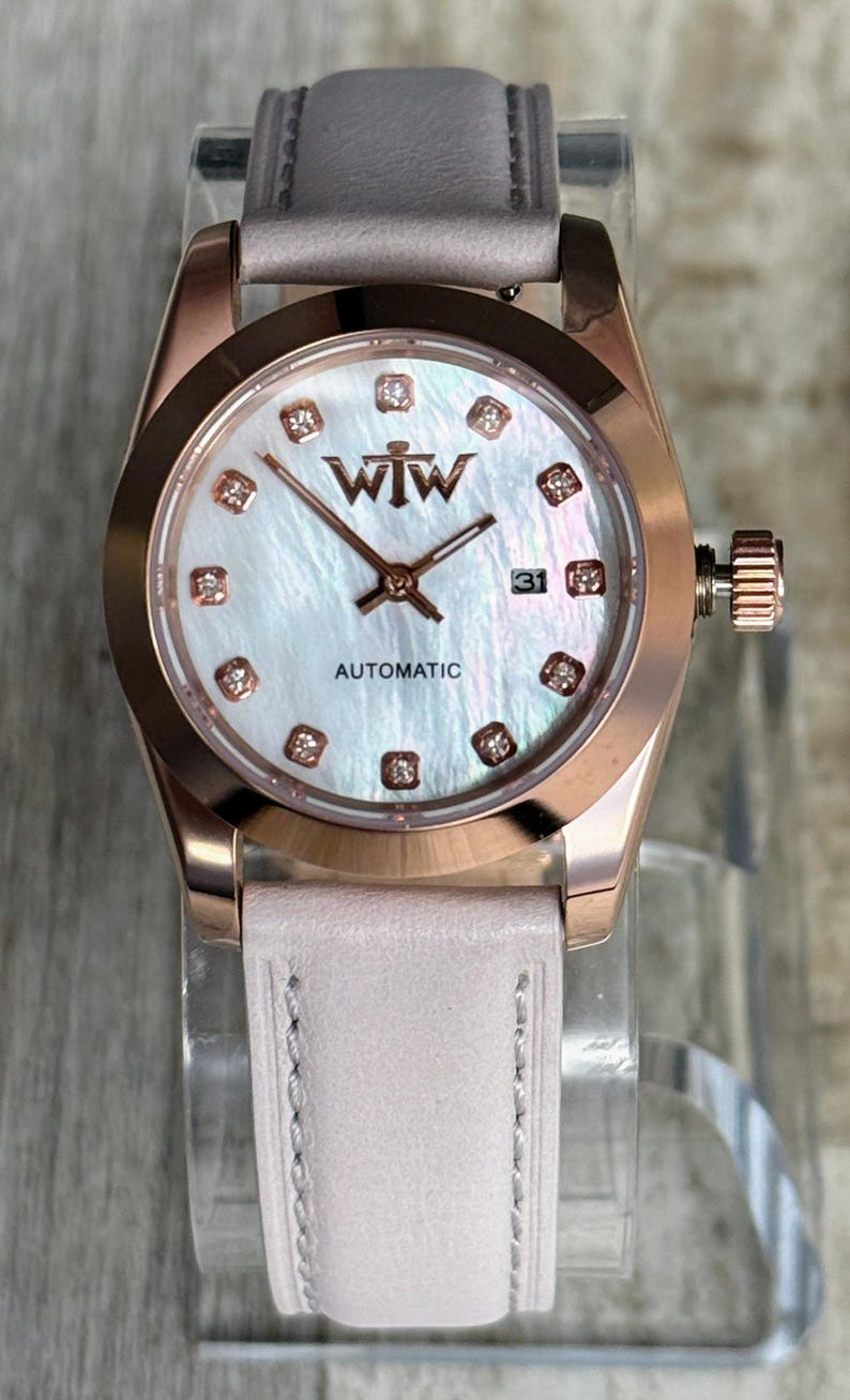 May include: A rose gold-tone wristwatch with a mother-of-pearl dial, featuring the text WTW and AUTOMATIC. The dial includes small crystal hour markers and a date window at the three o'clock position. It has a light pink leather strap.