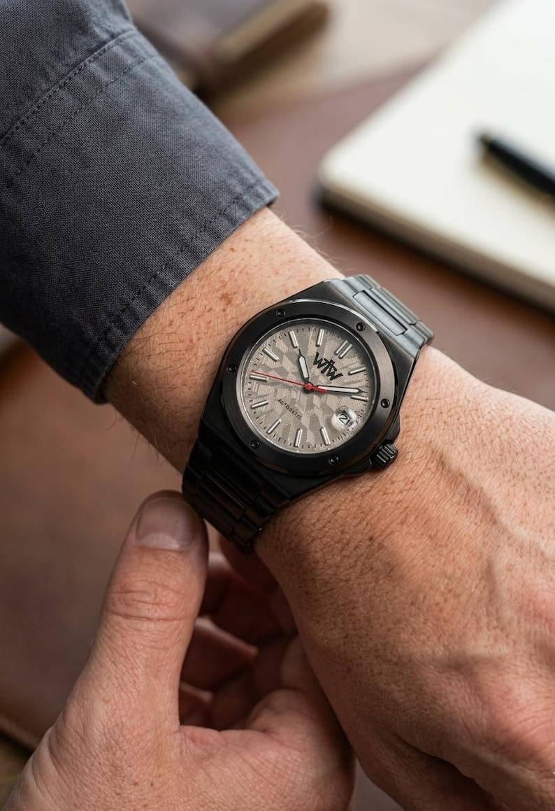 K&ouml;nnte beinhalten: Eine schwarze Armbanduhr aus Metall mit einem strukturierten hellgrauen Zifferblatt, dem Logo WtW, einem roten Sekundenzeiger und einem Datumsfenster auf der Drei-Uhr-Position. Die Uhr hat eine schwarze L&uuml;nette mit sichtbaren Schrauben und ein Gliederarmband.
