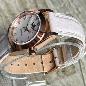 May include: An automatic wristwatch with a rose gold-tone case, a white mother-of-pearl dial featuring crystal hour markers, and the text WTW. The watch includes a white leather strap with a rose gold-tone buckle and a date display window.