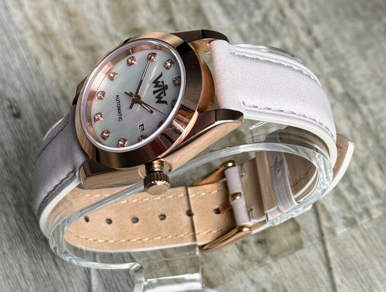 May include: An automatic wristwatch with a rose gold-tone case, a white mother-of-pearl dial featuring crystal hour markers, and the text WTW. The watch includes a white leather strap with a rose gold-tone buckle and a date display window.