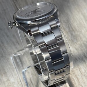 Puede incluir: Un reloj anal&oacute;gico de tono plateado con una esfera redonda de patr&oacute;n radial, manecillas plateadas y una corona situada en el lado izquierdo. El reloj incluye una pulsera de eslabones de acero inoxidable pulido y cepillado con cierre desplegable.