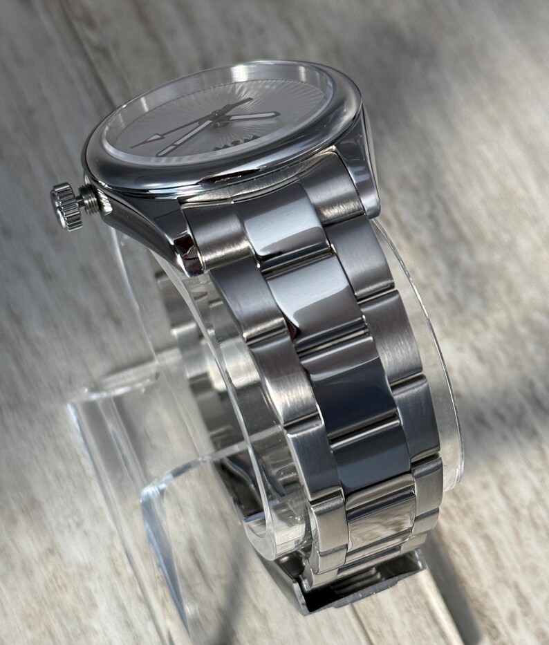 Puede incluir: Un reloj anal&oacute;gico de tono plateado con una esfera redonda de patr&oacute;n radial, manecillas plateadas y una corona situada en el lado izquierdo. El reloj incluye una pulsera de eslabones de acero inoxidable pulido y cepillado con cierre desplegable.