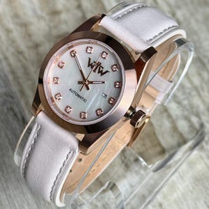 May include: An automatic wristwatch with a rose gold-tone case and a white leather strap. The watch face features a mother-of-pearl dial, rose gold hands, diamond-style hour markers, and the text WTW and AUTOMATIC with a date window at three.