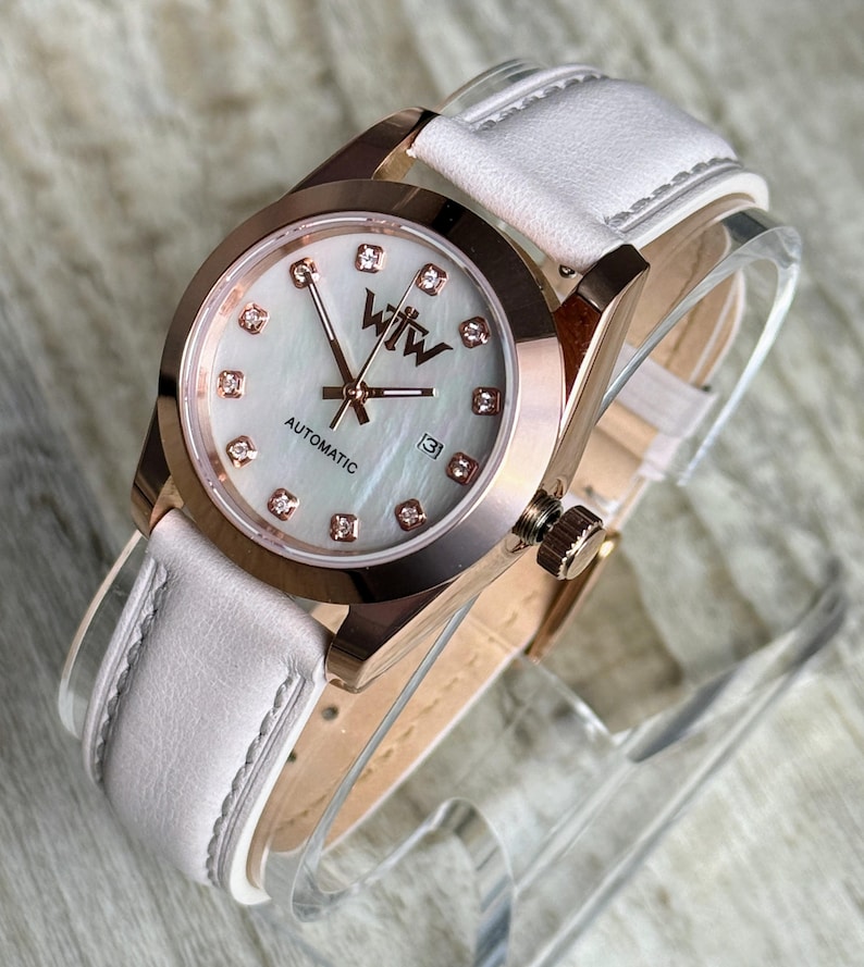 May include: An automatic wristwatch with a rose gold-tone case and a white leather strap. The watch face features a mother-of-pearl dial, rose gold hands, diamond-style hour markers, and the text WTW and AUTOMATIC with a date window at three.
