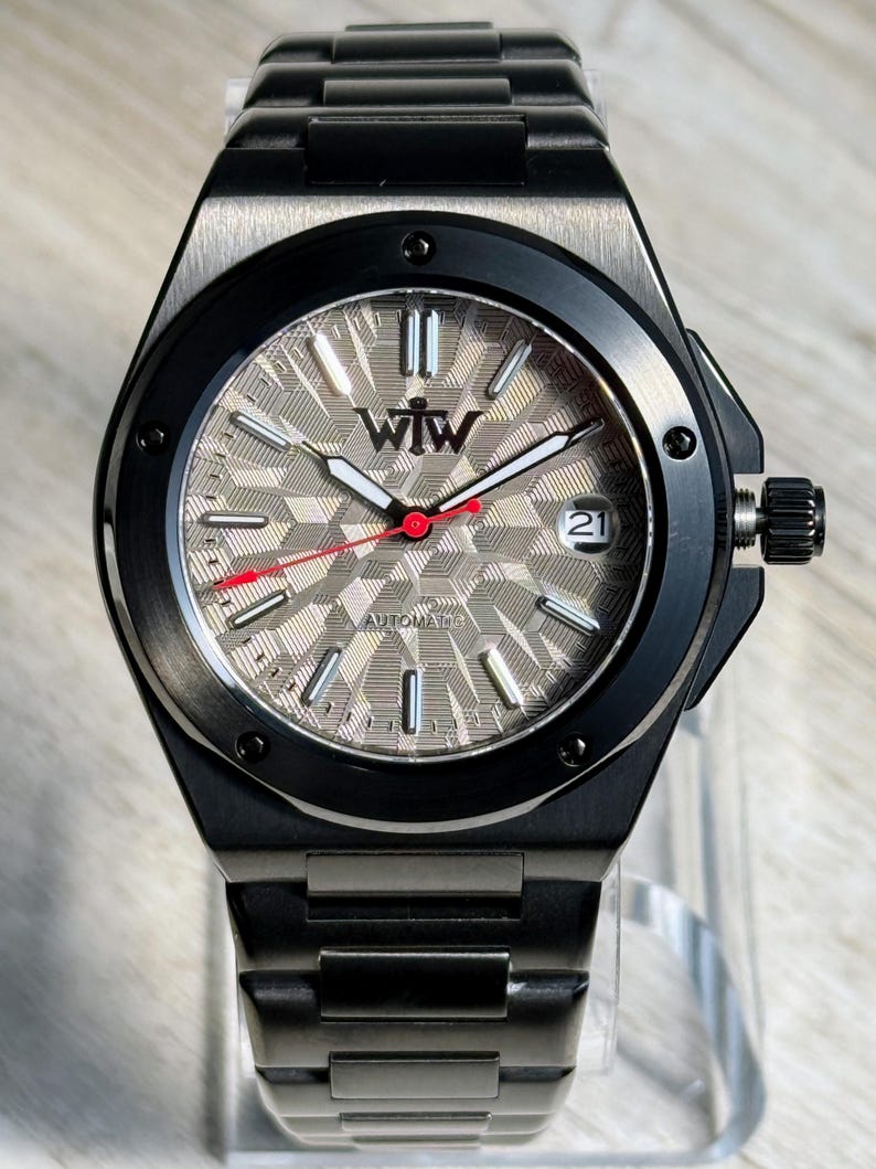 K&ouml;nnte beinhalten: Eine automatische Armbanduhr aus schwarzem Edelstahl mit einem strukturierten, silberfarbenen Zifferblatt mit geometrischem Muster. Das Zifferblatt zeigt das WTW-Logo, silberne Indizes, wei&szlig;e Leuchtzeiger, einen roten Sekundenzeiger und ein Datumsfenster mit der 21.