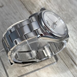 Puede incluir: Un reloj de pulsera plateado con una correa de eslabones de metal cepillado y una caja redonda. La esfera presenta un patr&oacute;n de rayos de sol, manecillas plateadas y el texto MVM en negro. Una peque&ntilde;a corona se sit&uacute;a en el lateral de la caja.