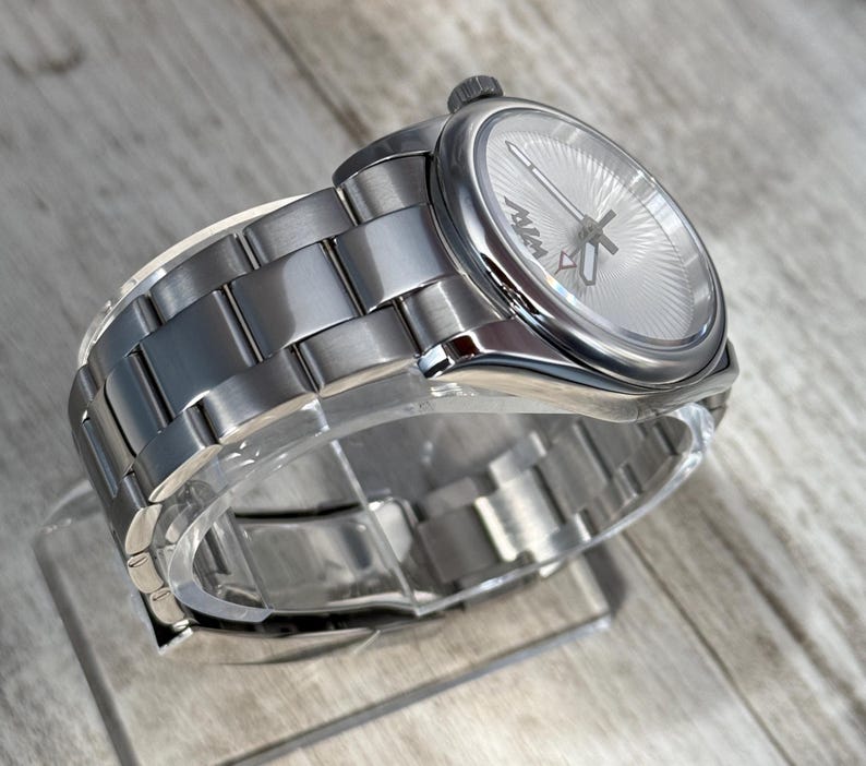 Puede incluir: Un reloj de pulsera plateado con una correa de eslabones de metal cepillado y una caja redonda. La esfera presenta un patr&oacute;n de rayos de sol, manecillas plateadas y el texto MVM en negro. Una peque&ntilde;a corona se sit&uacute;a en el lateral de la caja.