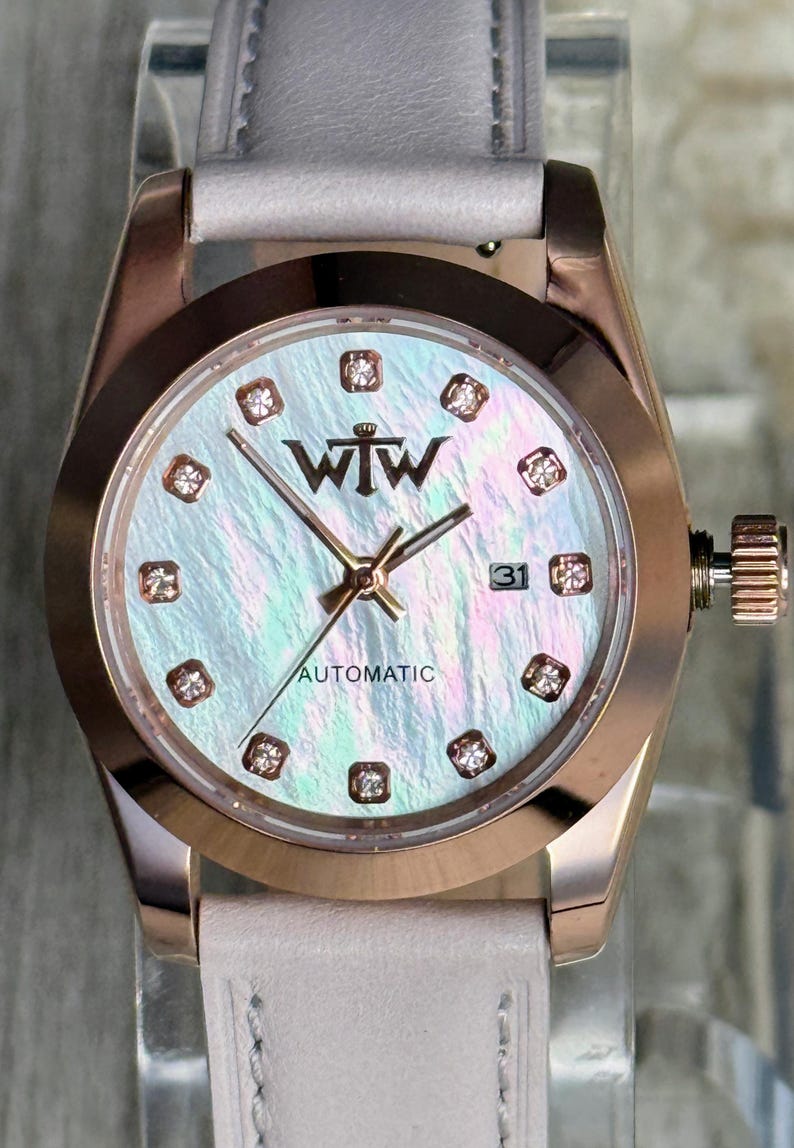 May include: A rose gold automatic watch with a light pink leather strap. The watch face features an iridescent mother-of-pearl dial, diamond-style hour markers, gold hands, and the text WTW and AUTOMATIC. A date window displays the number 31.