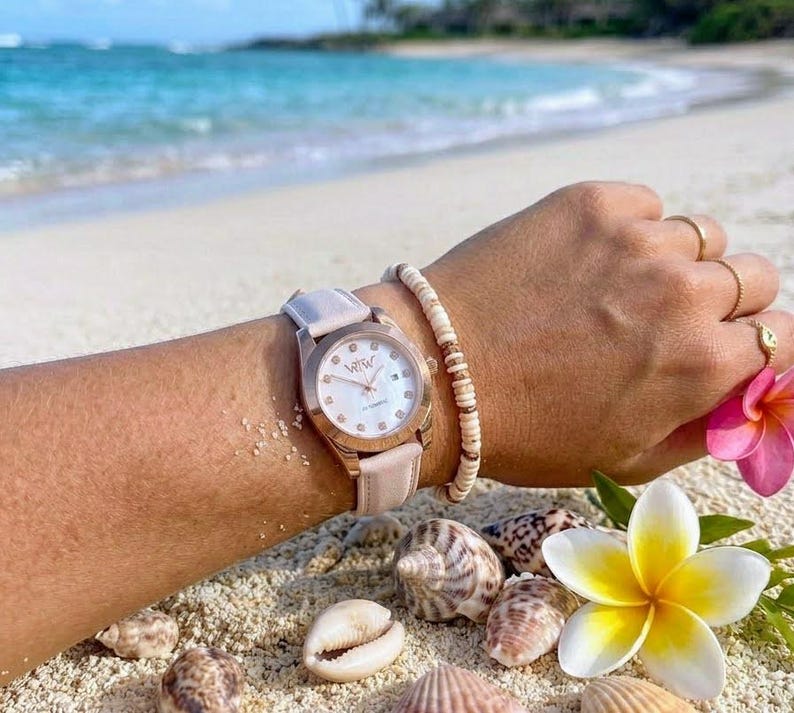 May include: A rose gold wristwatch with a light pink leather strap and a white dial featuring the letters WTW, crystal hour markers, and a date window. The watch is worn with a beaded bracelet and three gold rings on the fingers.