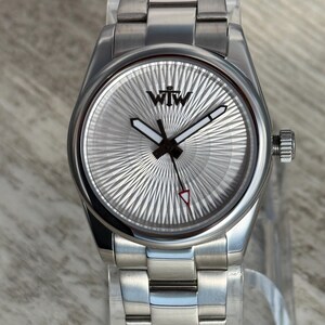 Puede incluir: Un reloj de pulsera de acero inoxidable en tono plateado con una esfera redonda con patr&oacute;n de rayos de sol y las letras WTW impresas en negro en la parte superior. El reloj incluye manecillas de hora y minutos con relleno blanco, un segundero fino con punta roja y una correa de eslabones.
