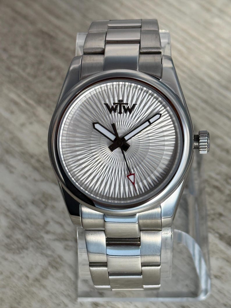 Puede incluir: Un reloj de pulsera de acero inoxidable en tono plateado con una esfera redonda con patr&oacute;n de rayos de sol y las letras WTW impresas en negro en la parte superior. El reloj incluye manecillas de hora y minutos con relleno blanco, un segundero fino con punta roja y una correa de eslabones.