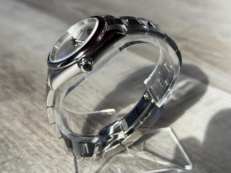 Puede incluir: Un reloj de pulsera anal&oacute;gico plateado con caja met&aacute;lica redonda y brazalete de eslabones a juego. La esfera tiene una textura plateada con manecillas plateadas y un peque&ntilde;o detalle rojo en el segundero, protegido por un cristal transparente.