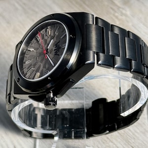 K&ouml;nnte beinhalten: Eine schwarze Automatikuhr mit einem matten Metallgliederarmband. Das Zifferblatt hat ein dunkelgraues, strukturiertes Muster, silberfarbene Stundenmarkierungen, wei&szlig;e Zeiger und einen roten Sekundenzeiger. Das Logo VTW ist auf dem Zifferblatt.